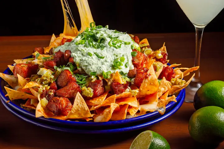 Recipe for homemade baked nacho chips topped with meat, melted cheese and sour cream with a glass of Margarita on the table